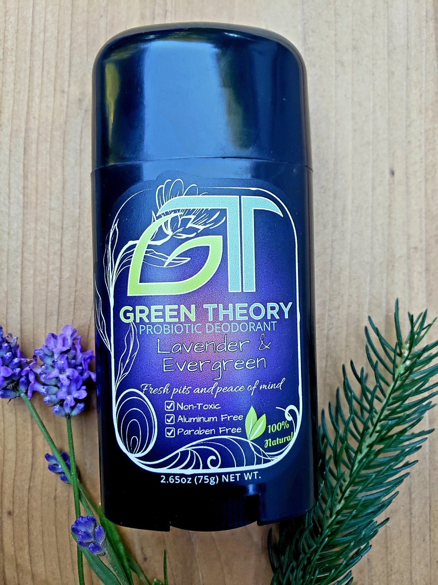 Photo of Green Theory Lavender & Evergreen womens deodorant. Deodorant is laying on wood texture with a sprig of lavender and evergreen cutting placed on the sides of the deodorant.