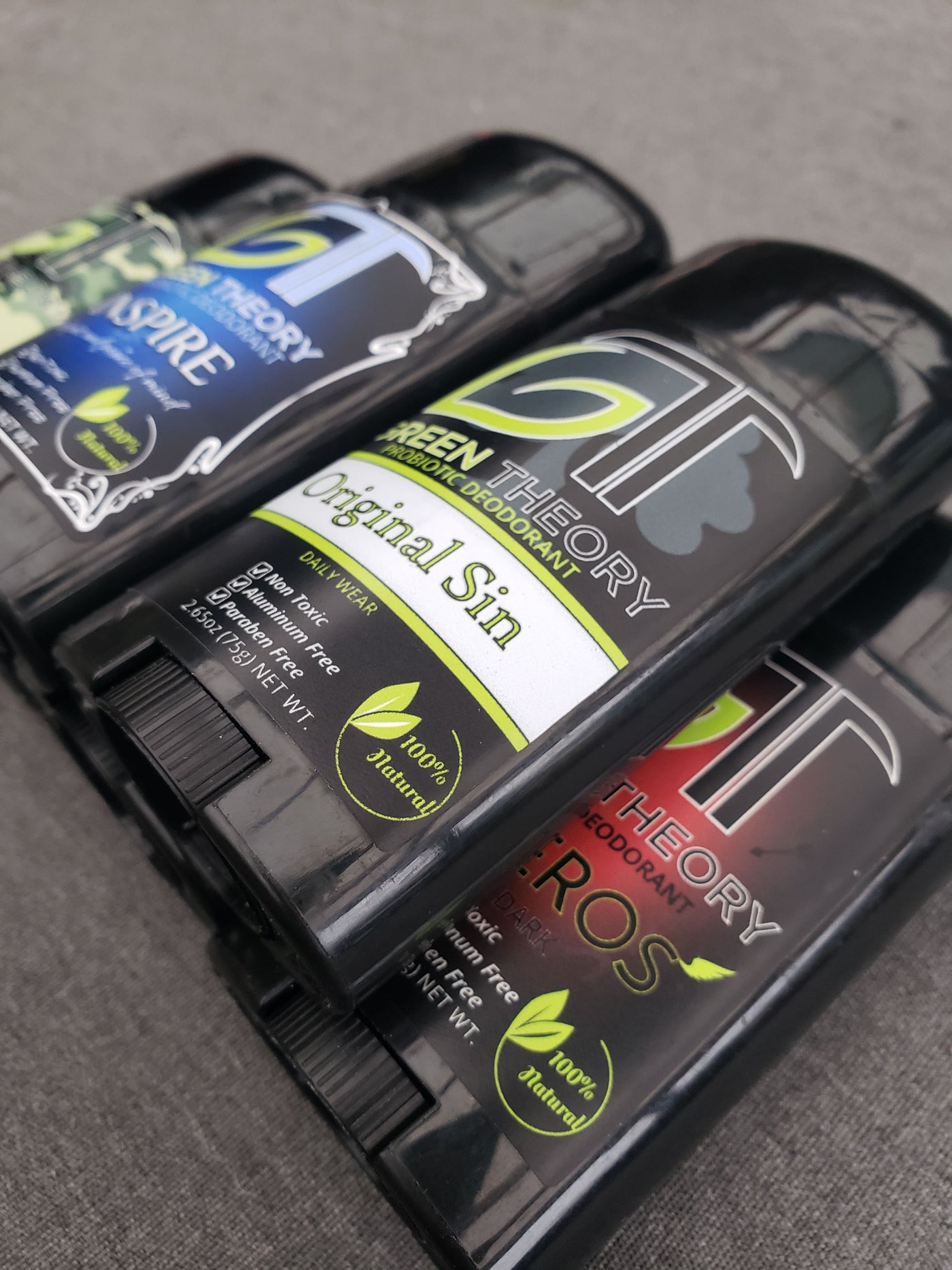 stack of green theory all natural probiotic deodorants. Pictured are original sin mens deodorant, inspire womens deodorant and anteros mens deodorant on a grey background. Deodorant sticks are a sleek black plastic container with large GT logo and scent