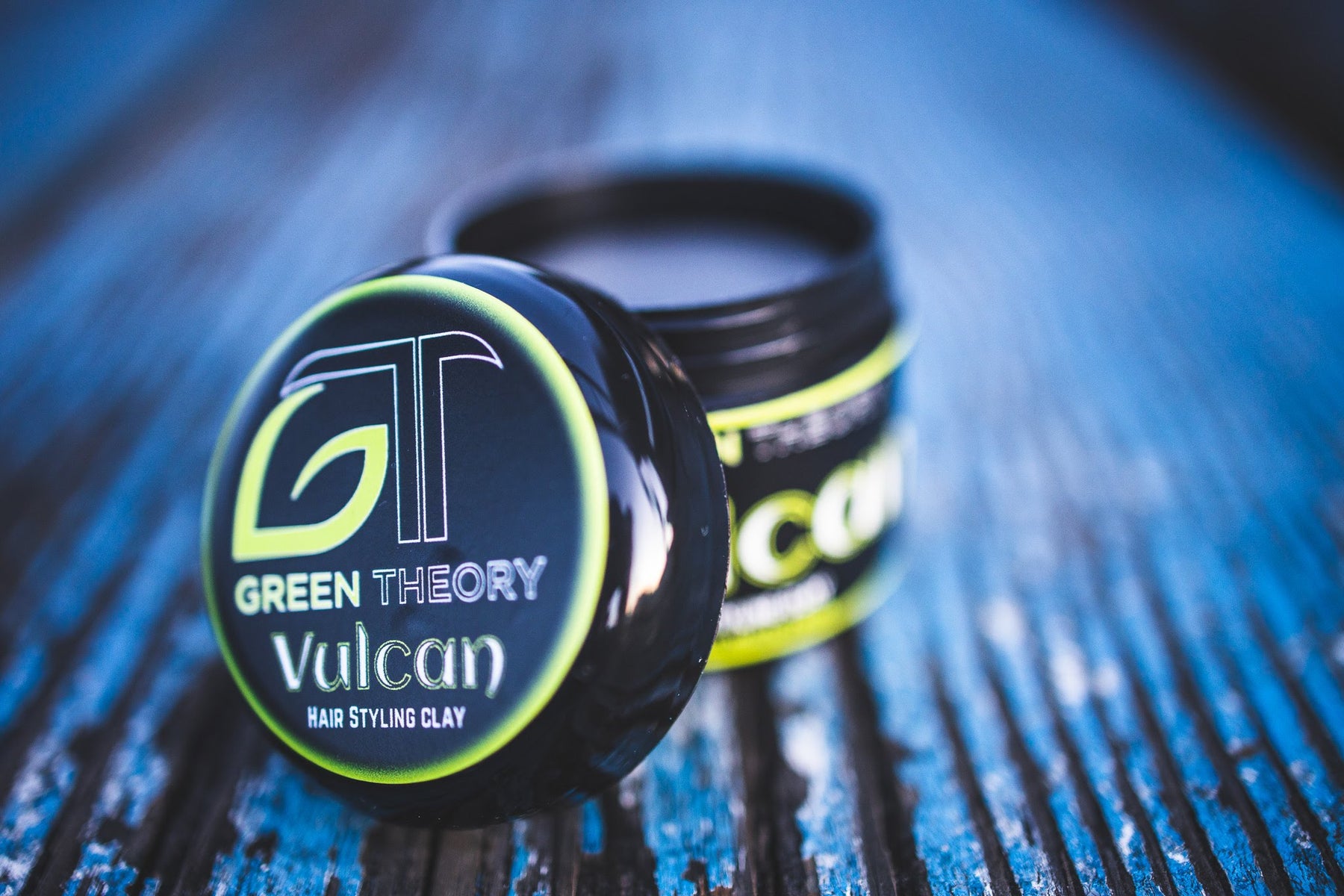 vulcan all natural hair styling clay with cap off. background is blurry, blue hued wood texture. Jar is black plastic with a label on the cap which is propped up against the opened jar of hair styling clay.