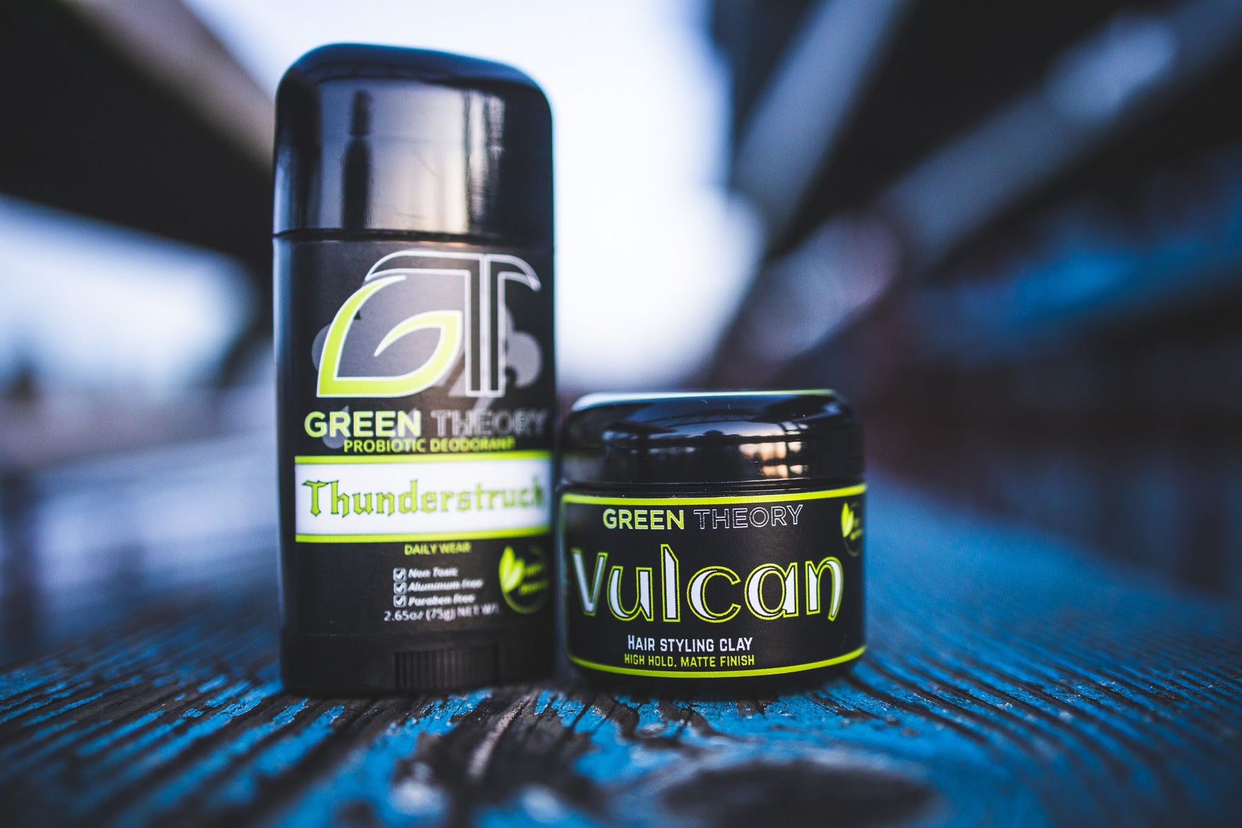 photograph of a stick of Green Theory's Thunderstuck probiotic, aluminum free deodorant and a jar of Vulcan hair styling clay sitting on a blue wood grain