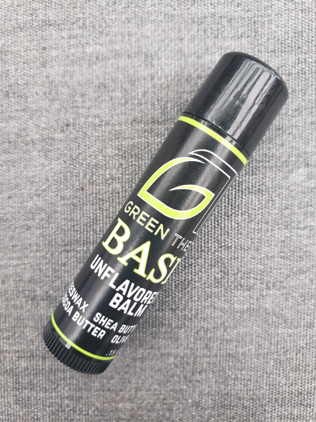 Close up photo of green theory basic unfloavored lip bal against a grey felt background. product is laying at an angle