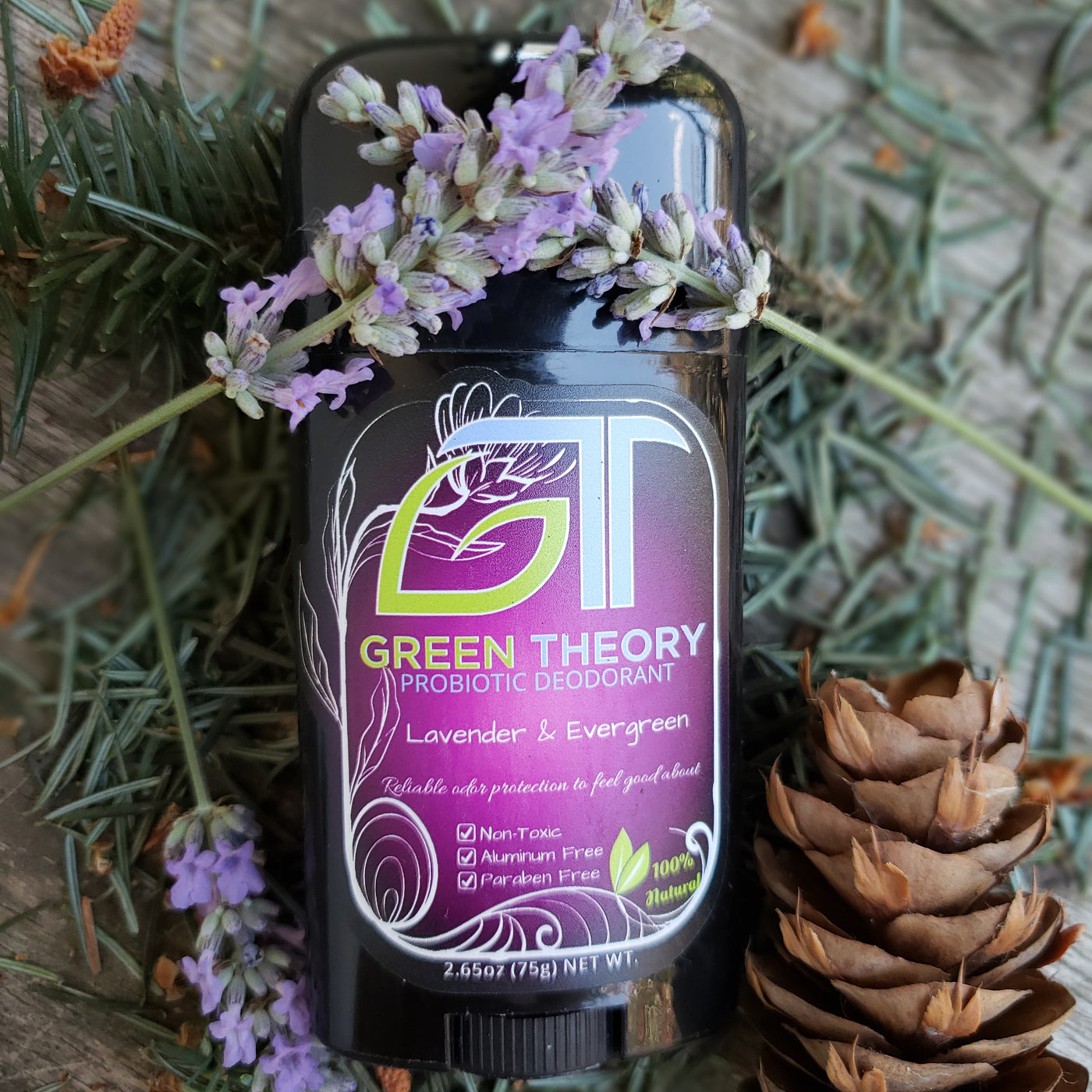 photo of green theory lavender $evergreen probiotic natural aluminum free deodorant for women laying with its front label up on top of a wood texture surrounded by pine needles, a pine cone and lavender sprigs