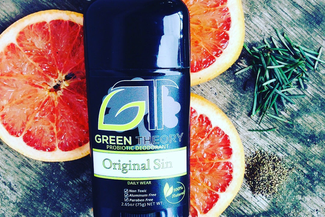 saturated image of a stick of green theory original sin probiotic deodorant for men laying on a wood grain background surrounded by slices of grapefruit, a pile of black pepper and fir needles to depict scent