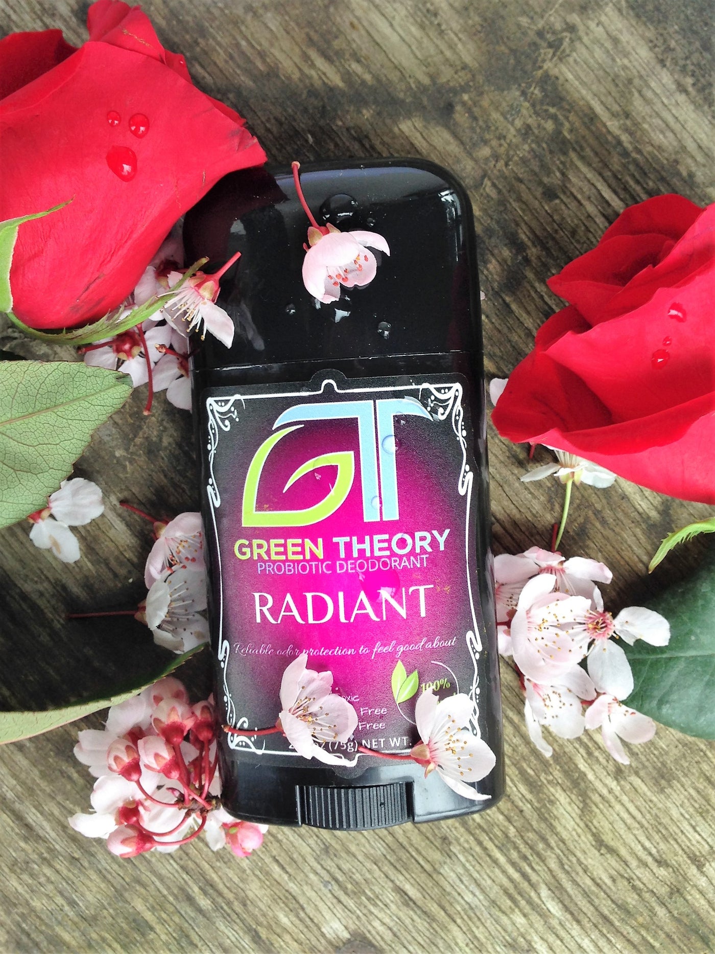 green theory radiant probiotic natural aluminum free deodorant laying on a wood grain texture with two roses placed on each side and little white flowers sprinkled over the container