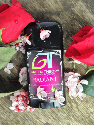 green theory radiant probiotic natural aluminum free deodorant laying on a wood grain texture with two roses placed on each side and little white flowers sprinkled over the container