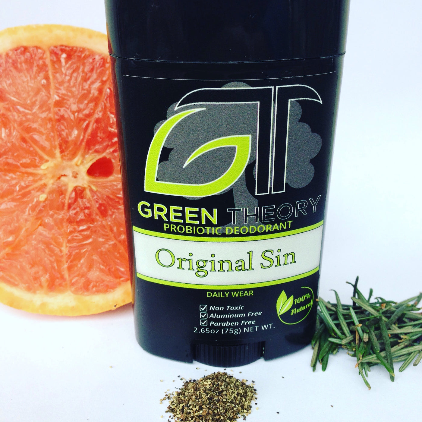 photo looking down at the front of a stick of green theory original sin probiotic natural deodorant for men. background is pure white and the stick is surrounded by a grapefruit slice, black pepper and fir needles to illustrate the scent